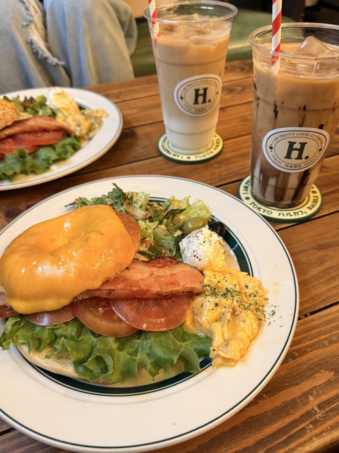 H.ロゴ入りプラスチックカップのアイスラテ2杯と、チェダーチーズが溶けたベーグルに厚切りベーコン・トマト・レタス・スクランブルエッグ・ポーチドエッグが盛られたプレート